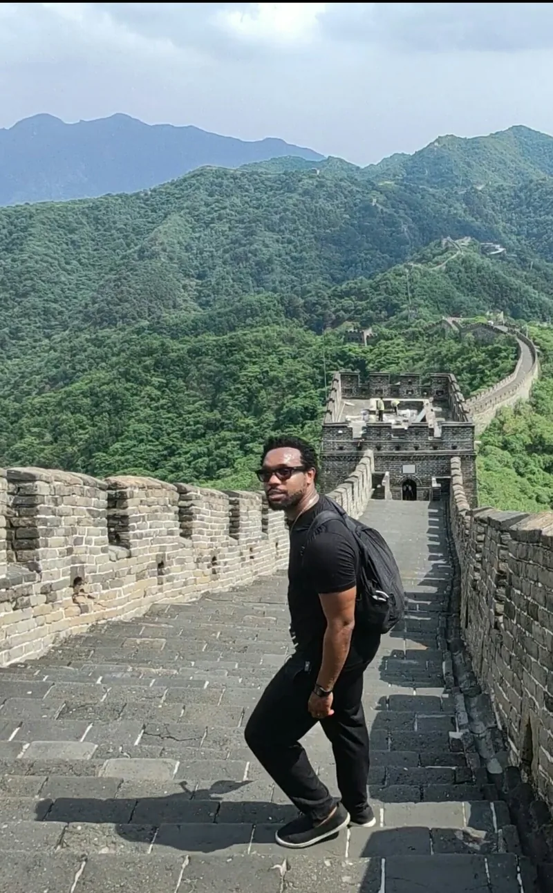 Bakari Akil at the Great Wall of China Bakari Akil climbing the Great Wall of China in sunglasses and a black polo, looking back at the camera with green mountains and watchtowers stretching into the distance behind him