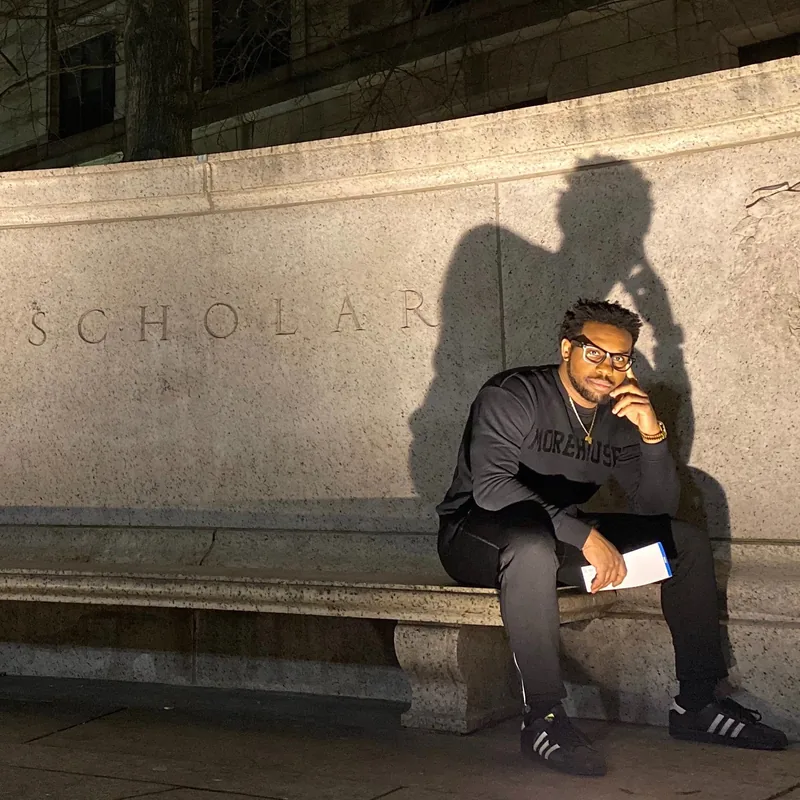 Bakari Akil, author of Paper Ceiling, reading beneath SCHOLAR inscription Bakari Akil sitting on a stone bench beneath the word SCHOLAR carved into a university wall, wearing a Morehouse College sweatshirt and glasses, reading a book at night — author photo for his forthcoming memoir Paper Ceiling