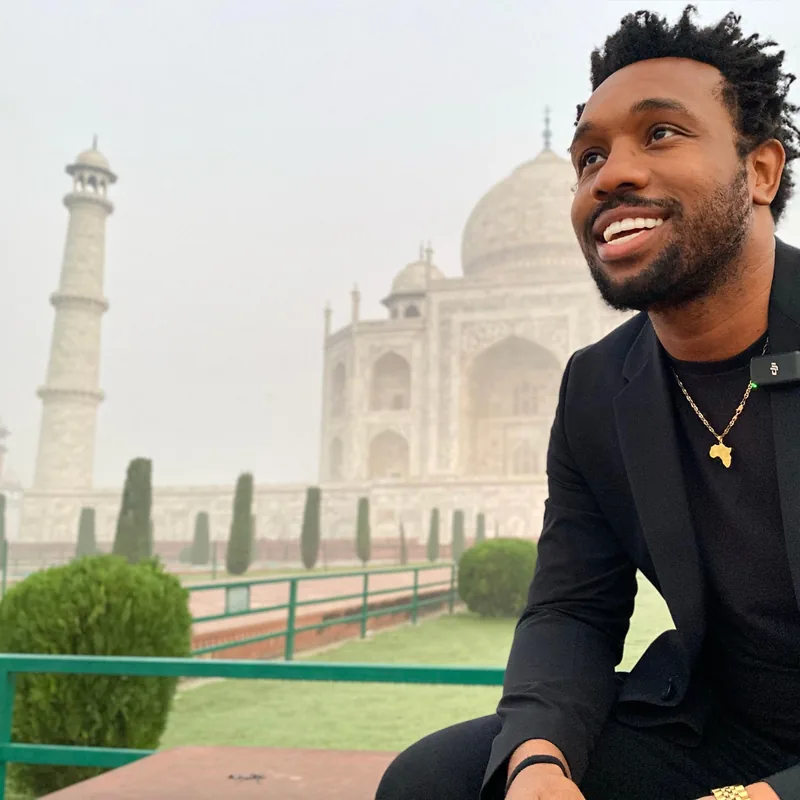 Bakari Akil at the Taj Mahal, Agra, India Bakari Akil smiling in a black blazer and gold Africa pendant necklace seated in front of the Taj Mahal in Agra, India, the white marble dome and minarets hazy in the morning light behind him