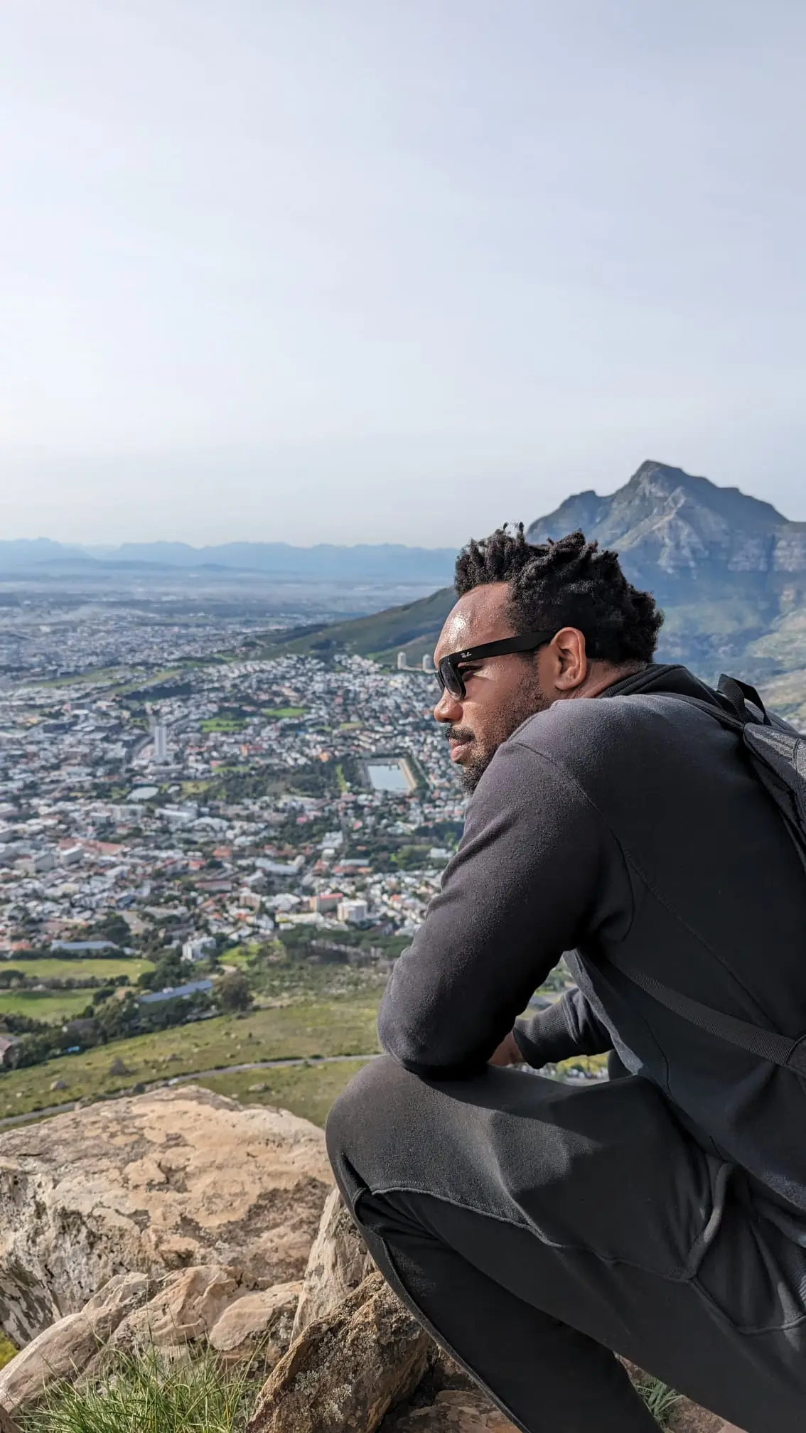 Bakari Akil hiking above Cape Town, South Africa Bakari Akil seated on a rocky summit overlooking the city of Cape Town, South Africa, wearing sunglasses and a black sweater with a backpack, Table Mountain and the Atlantic coastline stretching out below