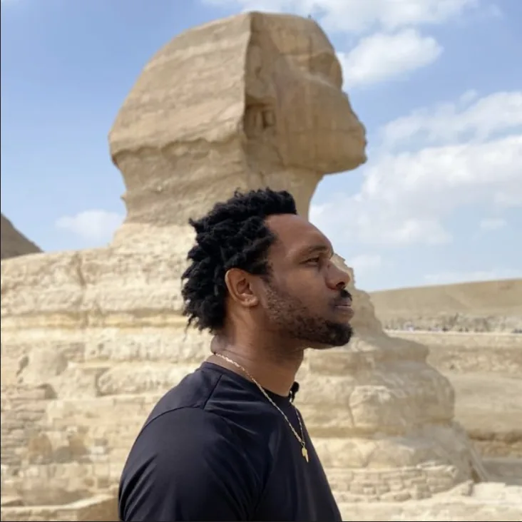 Bakari Akil at the Great Sphinx, Giza, Egypt Bakari Akil in profile beside the Great Sphinx of Giza, Egypt — his silhouette mirroring the Sphinx's, wearing a black t-shirt and gold chain with the Giza pyramids and blue sky behind him