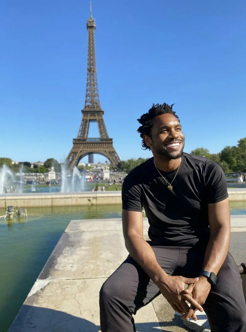 Bakari Akil at the Trocadéro with the Eiffel Tower, Paris, France Bakari Akil seated at the Trocadéro fountains in Paris smiling in a black t-shirt and gold chain, the Eiffel Tower rising behind him against a clear blue sky