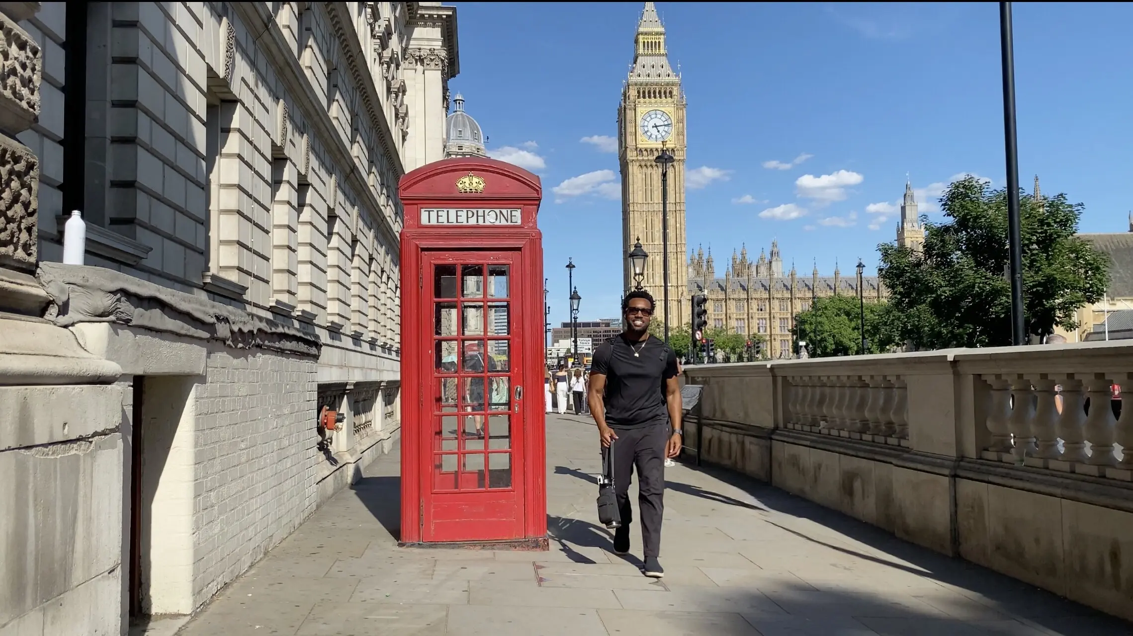 Bakari Akil at Big Ben and Westminster, London, England Bakari Akil walking past a red telephone box on Westminster Bridge in London, England, with Big Ben and the Houses of Parliament rising behind him under a blue sky