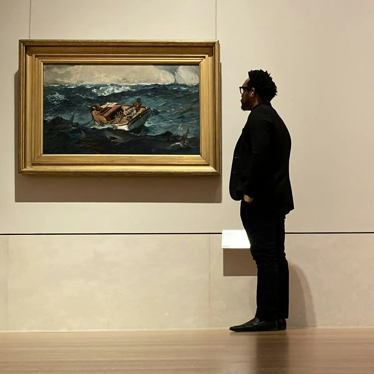 Bakari Akil at the Metropolitan Museum of Art, New York Bakari Akil, private equity investor and business acquisition lecturer, standing in profile in a museum gallery studying Winslow Homer's The Gulf Stream — a painting of a Black man alone on a broken boat surrounded by sharks — wearing all black with glasses and a gold watch