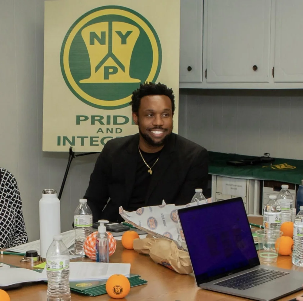 Bakari Akil at NYP Corp, a Graves Hall Capital portfolio company Bakari Akil smiling in the NYP Corp conference room, seated at a long table with a laptop open, wearing a black blazer and gold Africa pendant necklace, the green-and-gold NYP Pride and Integrity logo banner behind him — the $30M specialty manufacturer he acquired through Graves Hall Capital