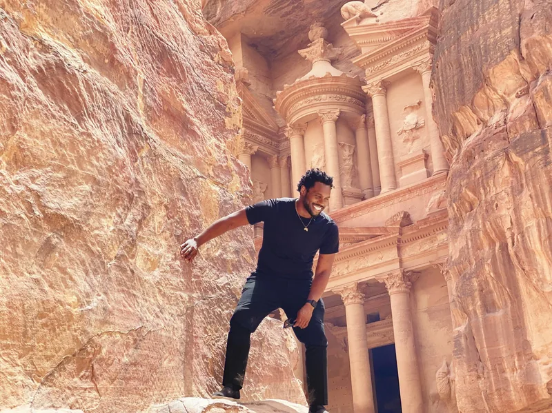 Bakari Akil at The Treasury (Al-Khazneh), Petra, Jordan Bakari Akil posing on a rock ledge in front of Al-Khazneh, the ancient Treasury carved into rose-red sandstone cliffs at Petra, Jordan, wearing all black