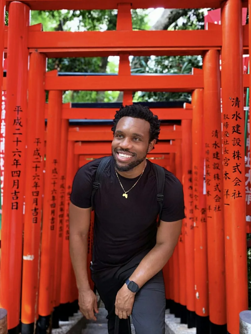 Bakari Akil at a torii gate shrine in Tokyo, Japan Bakari Akil laughing under a corridor of vermilion torii gates at a Shinto shrine in Tokyo, Japan, wearing a black t-shirt with a gold Africa pendant, Japanese kanji inscriptions visible on the gate pillars