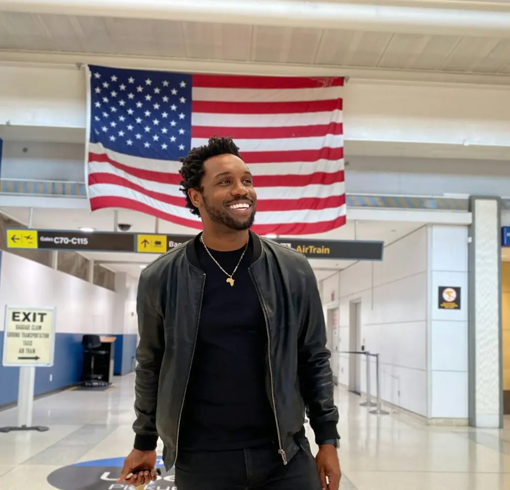 Bakari Akil at JFK International Airport, New York Bakari Akil smiling in a black leather bomber jacket and gold Africa pendant necklace walking through JFK Airport with a large American flag hanging overhead, Gates C70-C115 and AirTrain signs visible